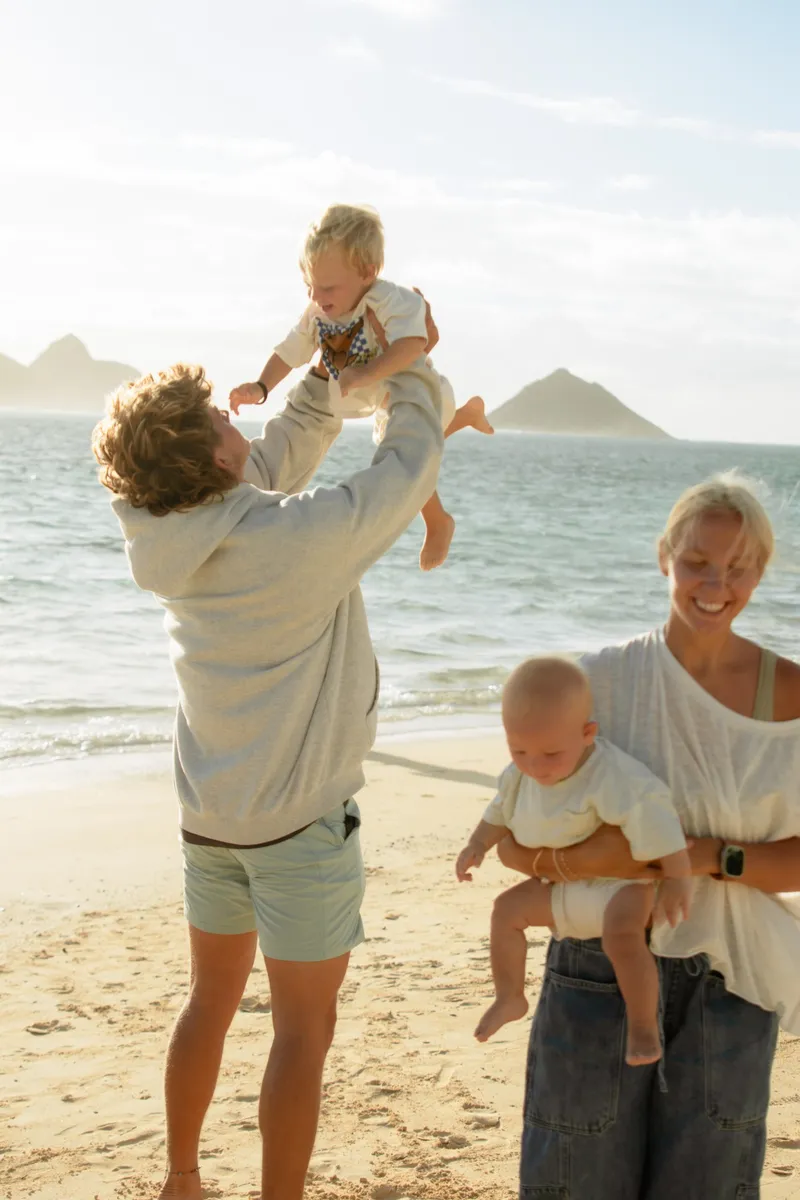Clay tossing their older son in the air while Ali holds their baby on the beach