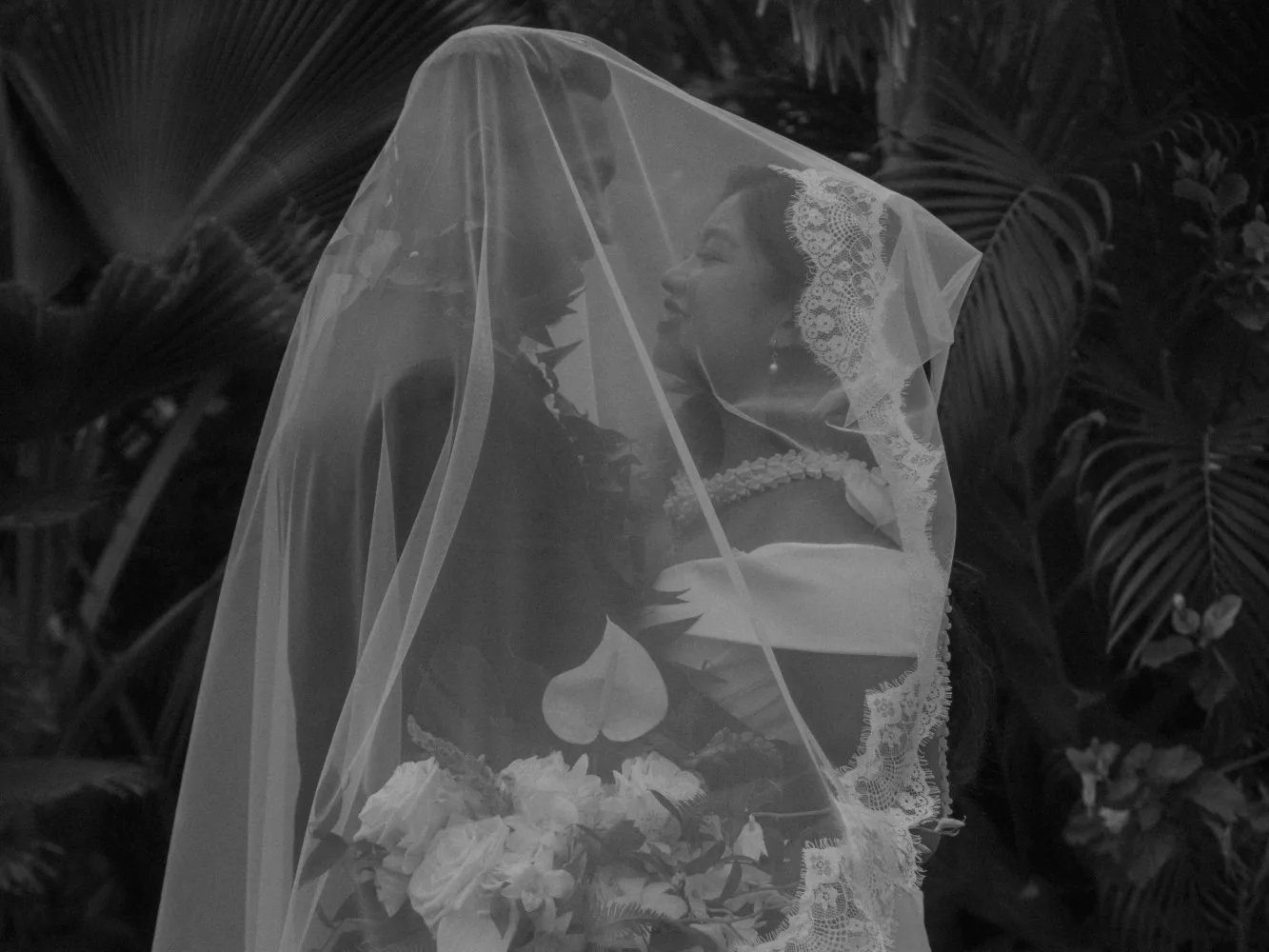 A bride and groom beneath her lace-edged veil, in black and white